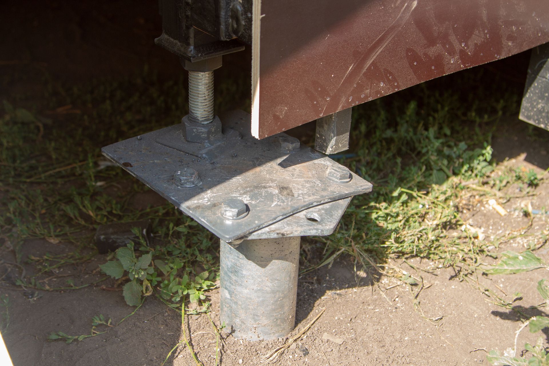Metal post with a square base plate and bolts on dirt beside a wooden structure.