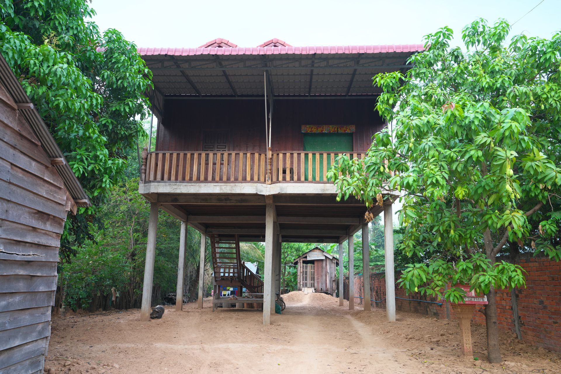 Wooden stilt house with front porch, surrounded by lush green trees on a dirt yard.