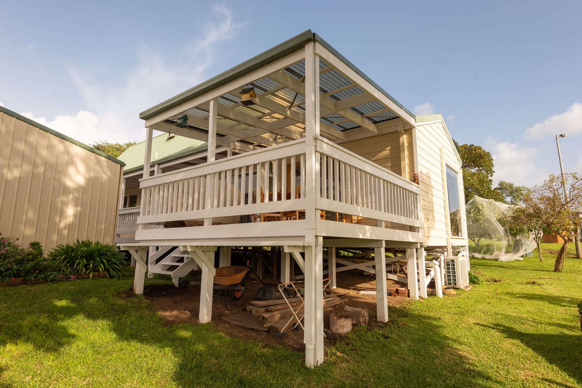 Raised white house on stilts with a porch, surrounded by grass and trees under a blue sky