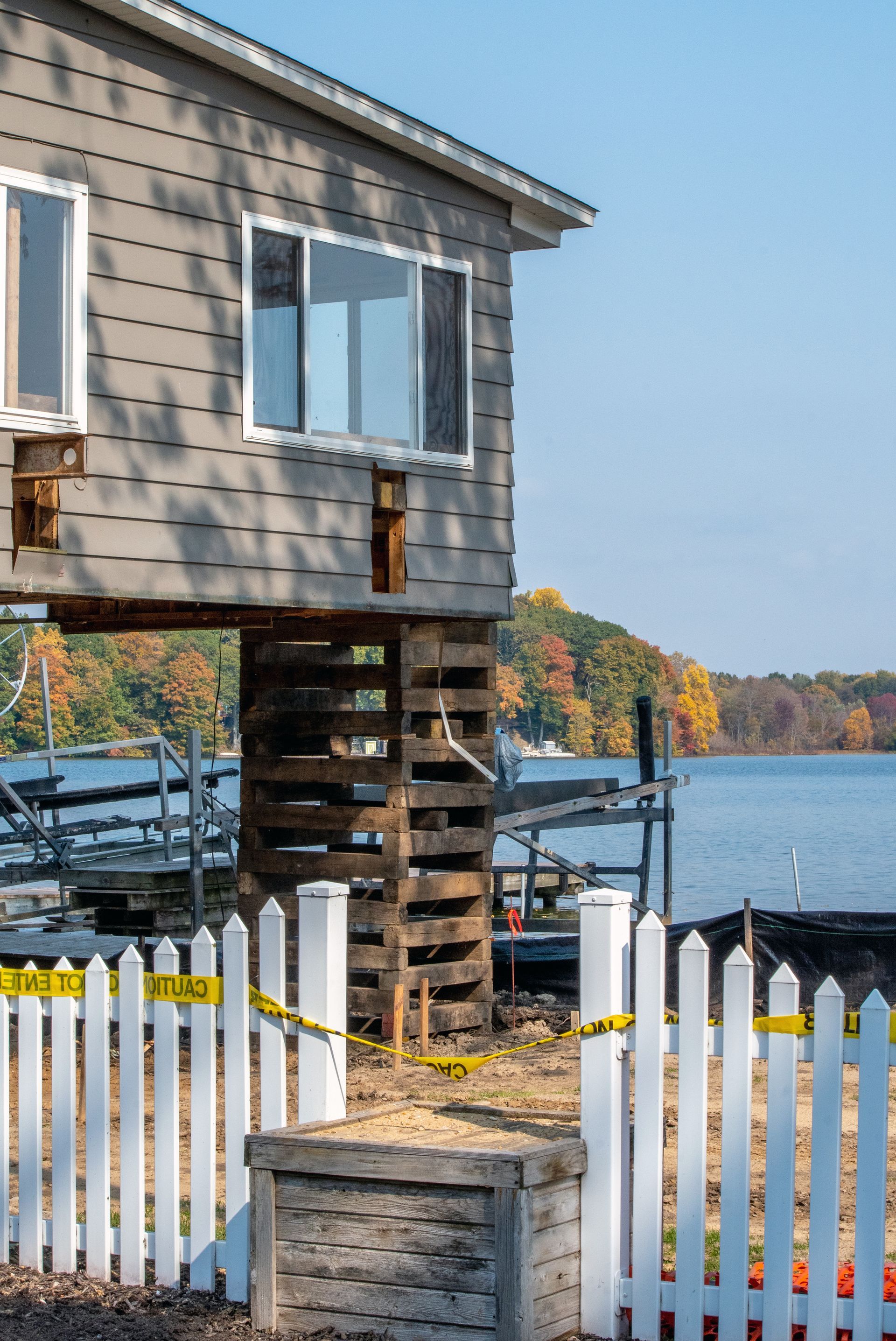 Elevated gray house on pilings by a calm waterfront with white fence and autumn trees beyond