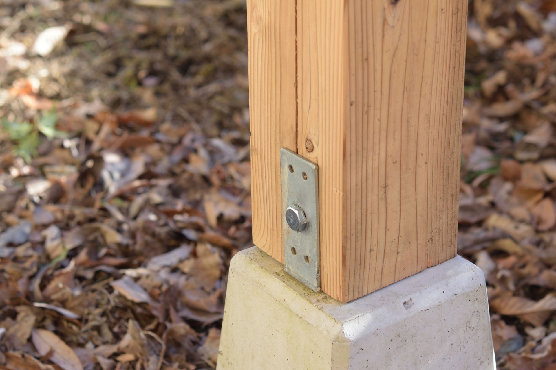 Wooden post bolted to a concrete base with a metal bracket outdoors on leaf-covered ground