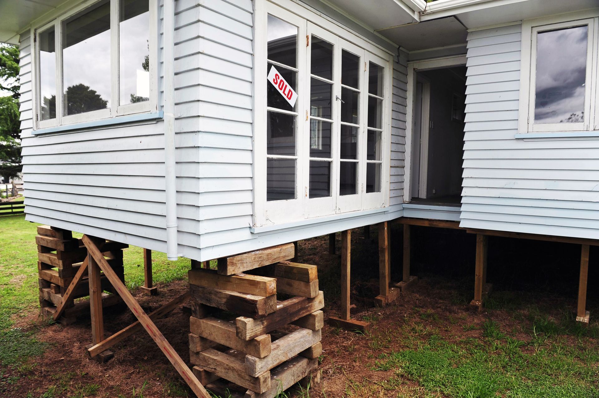 House elevated on stacked wooden supports with unfinished stairs and crawlspace visible underneath