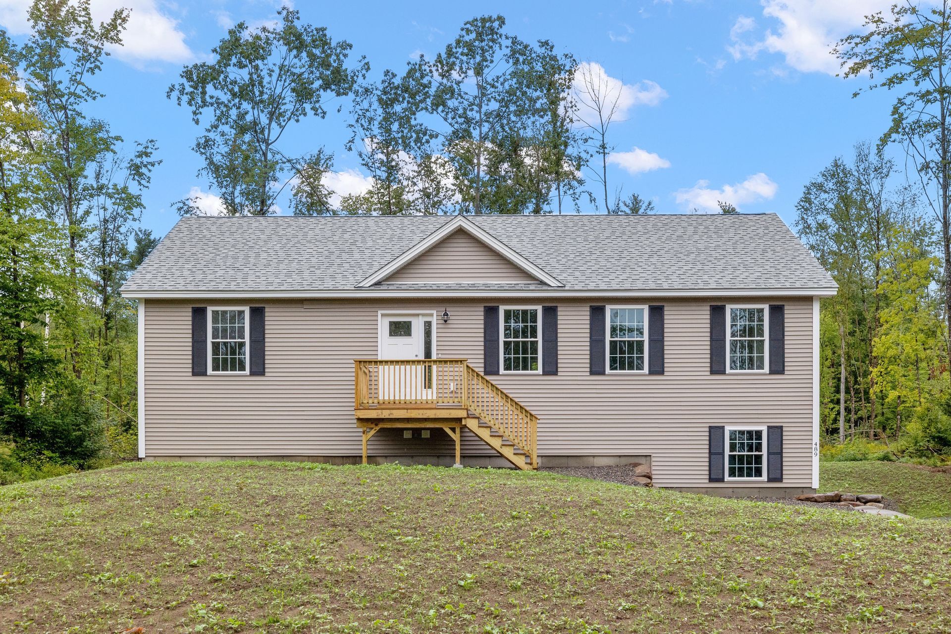 Single-story beige house with a small wooden porch, set on a grassy lot with trees and a blue sky.