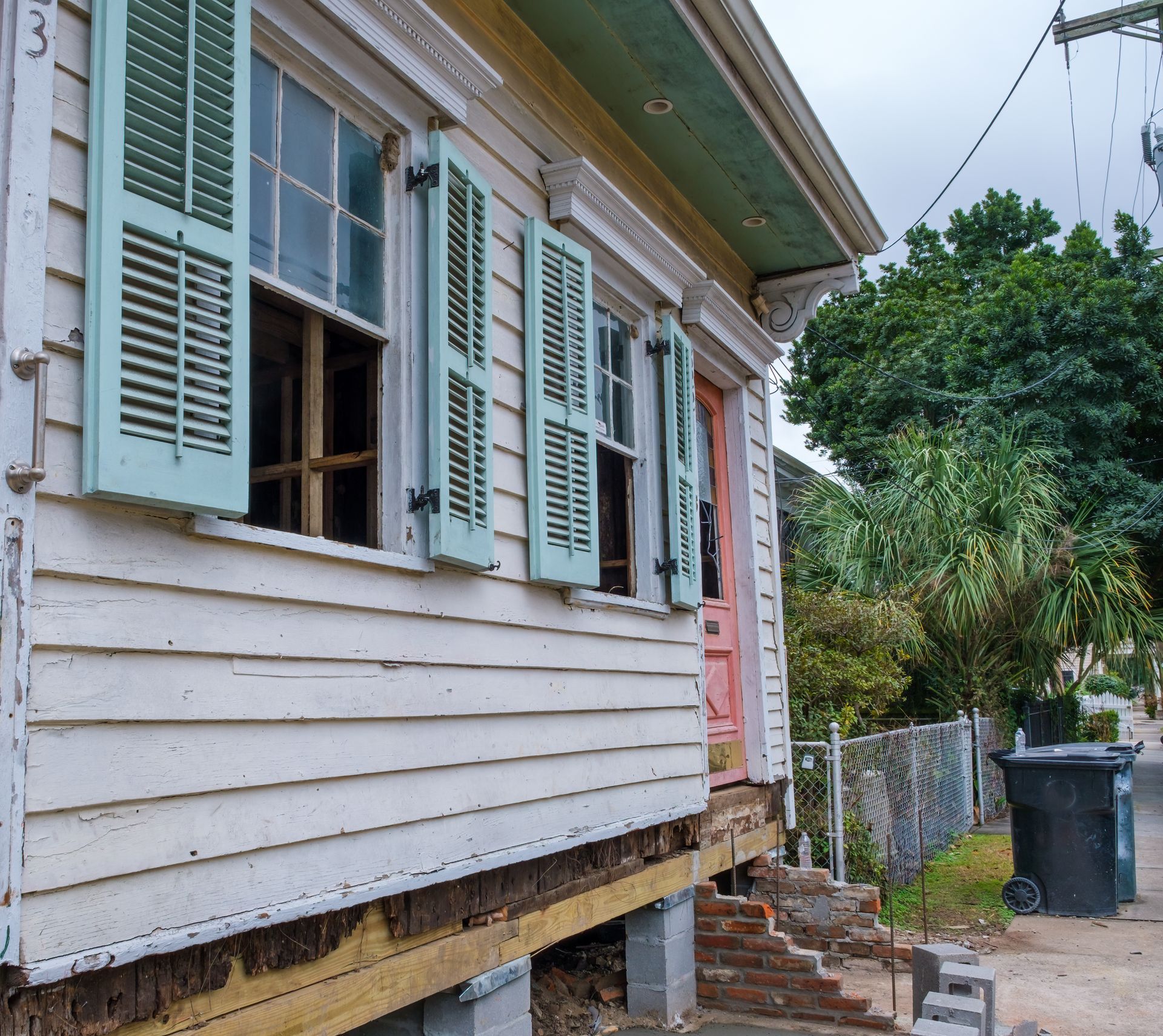 Weathered white house with mint shutters, raised foundation, and a side yard with trash bins and trees
