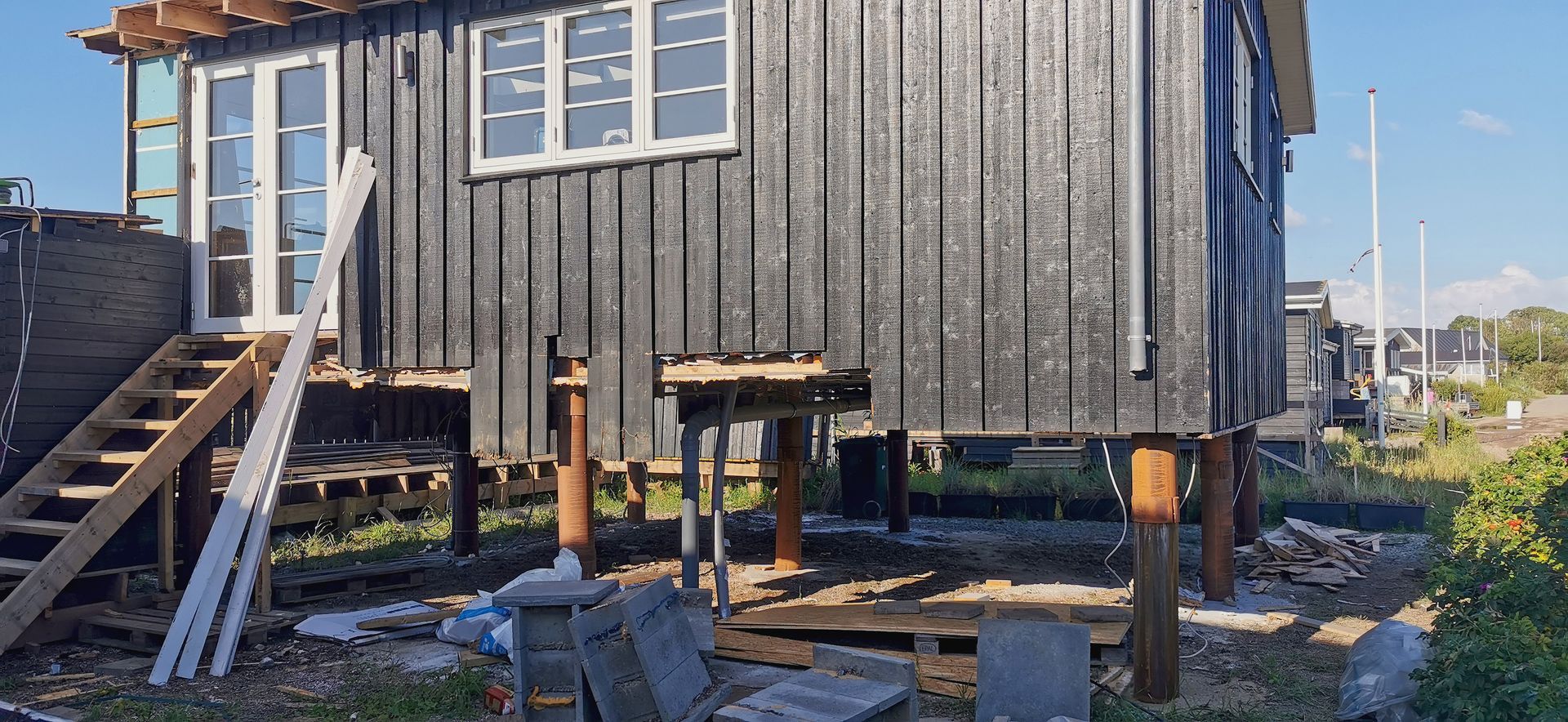 Elevated wooden house with stairs and supporting posts, shown in a sunny yard.