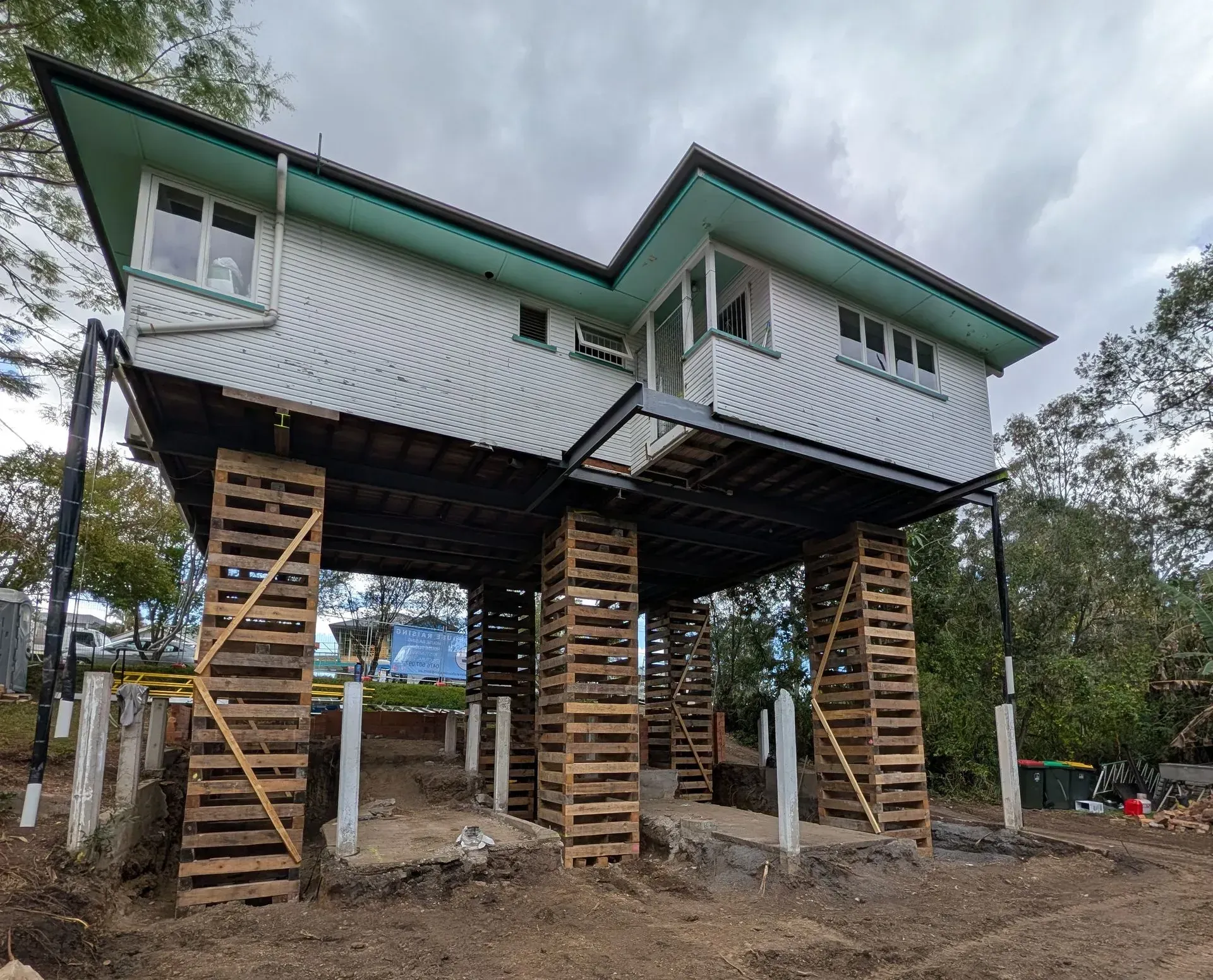 Elevated house on stilts with green roof and wooden access stairs, under construction on a muddy lot.