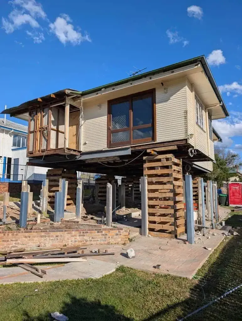 Elevated house under construction on stilts, with exposed framing and wooden scaffolding in a sunny yard.