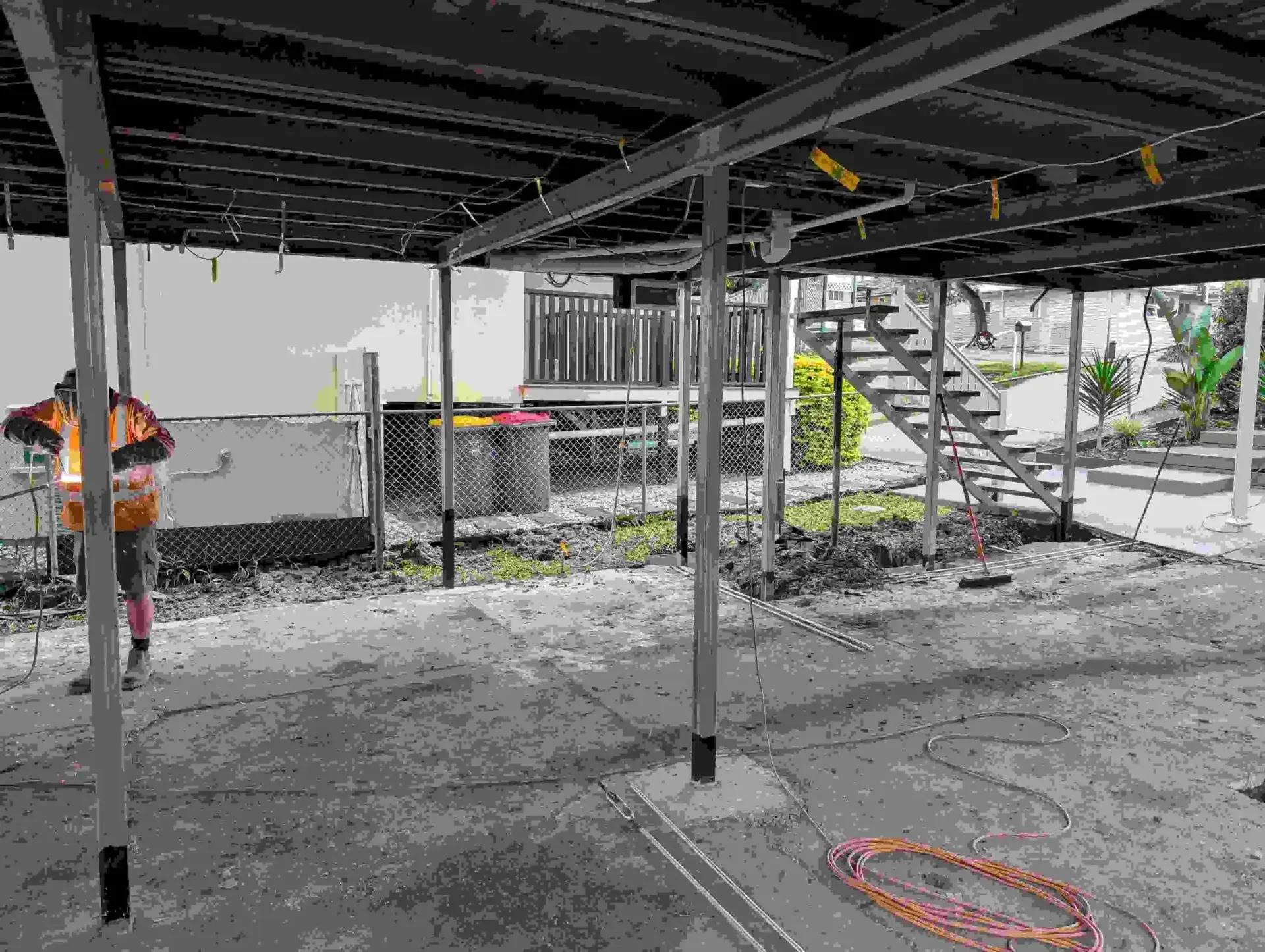 Construction site under a raised structure with steel beams, stairs, scaffolding, and gravel ground with orange hose.
