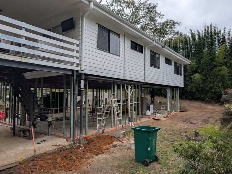 Raised house under construction on stilts, with scaffolding, dirt yard, and green trash bin.