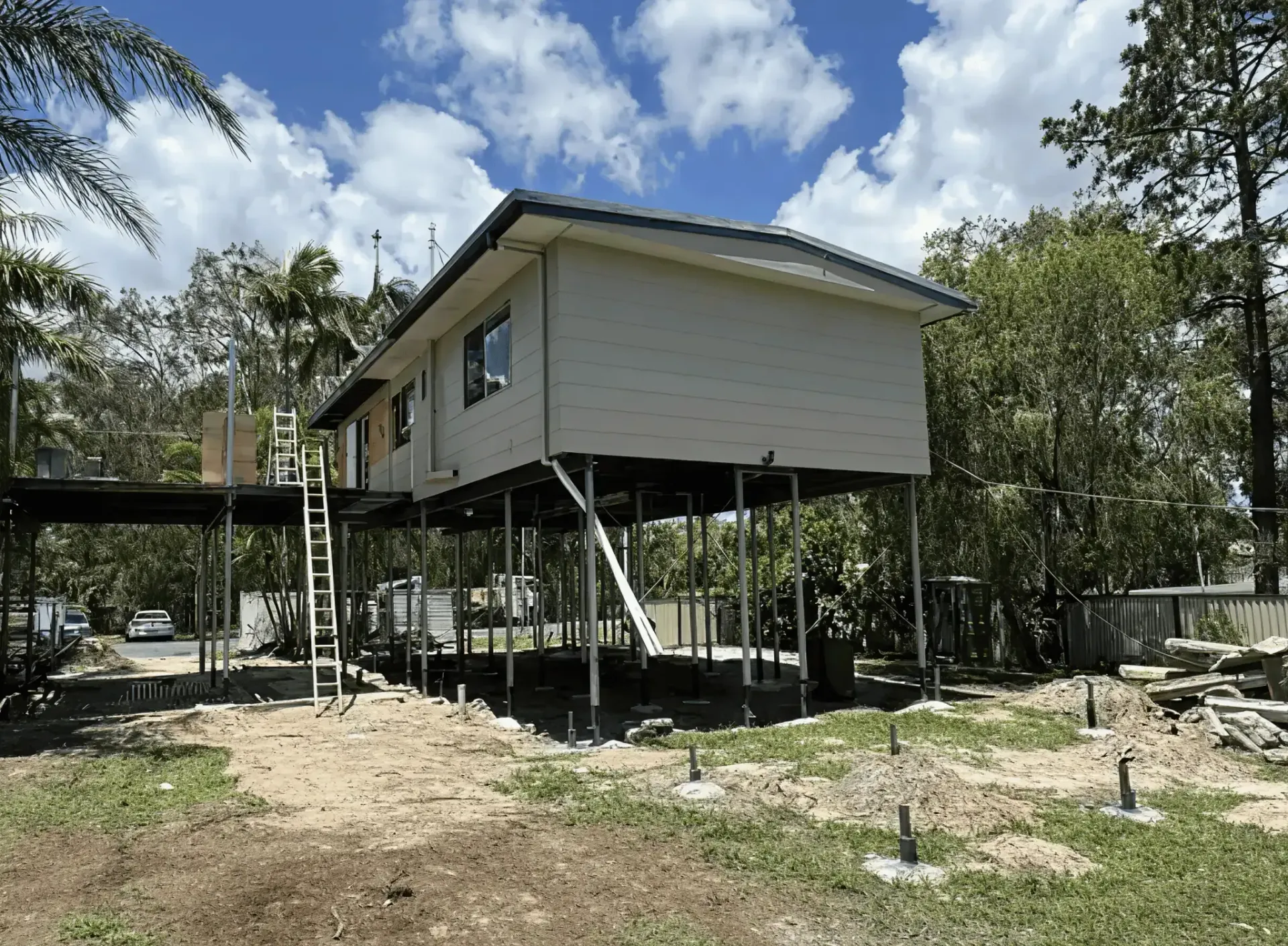 Elevated white house on stilts in a wooded area, with a covered walkway and rocky ground below.