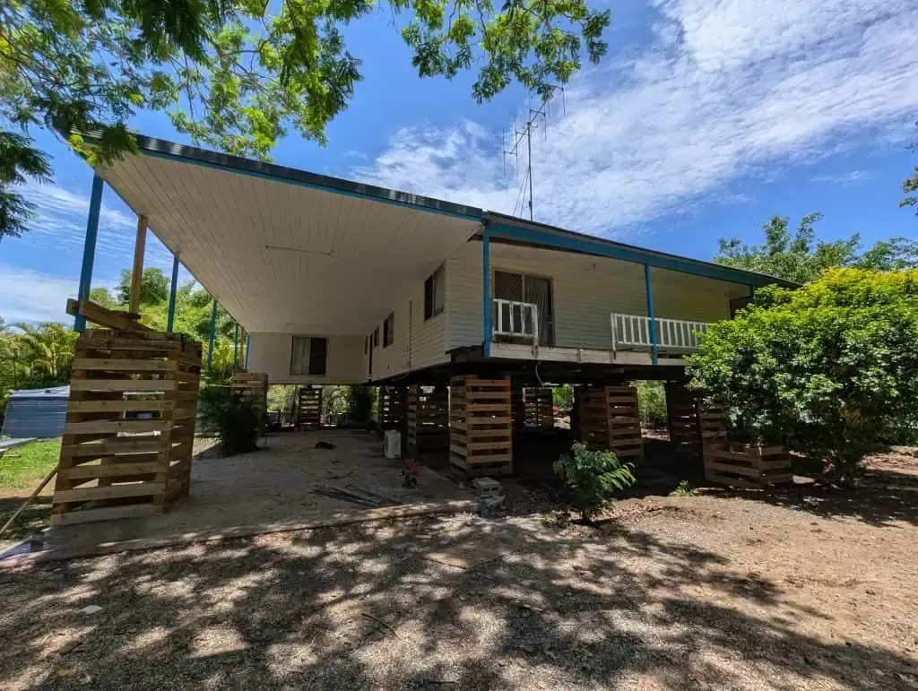 Modern elevated house with large slanted roof, wooden stairs, and surrounding greenery under a blue sky