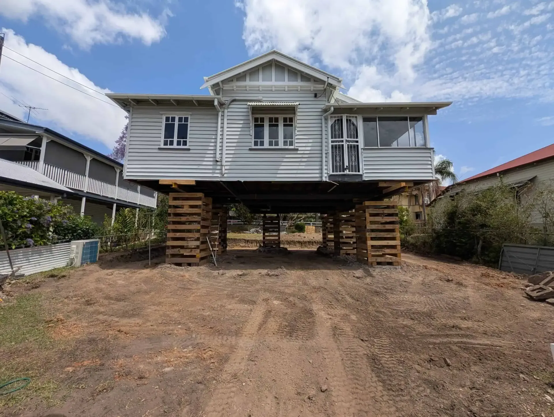 Raised white house on tall wooden stilts in a dirt yard under a blue sky