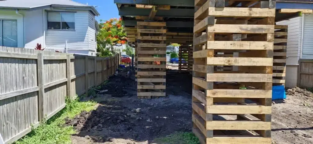 Outdoor area under a raised deck with stacked wooden support posts beside a white fence and house.