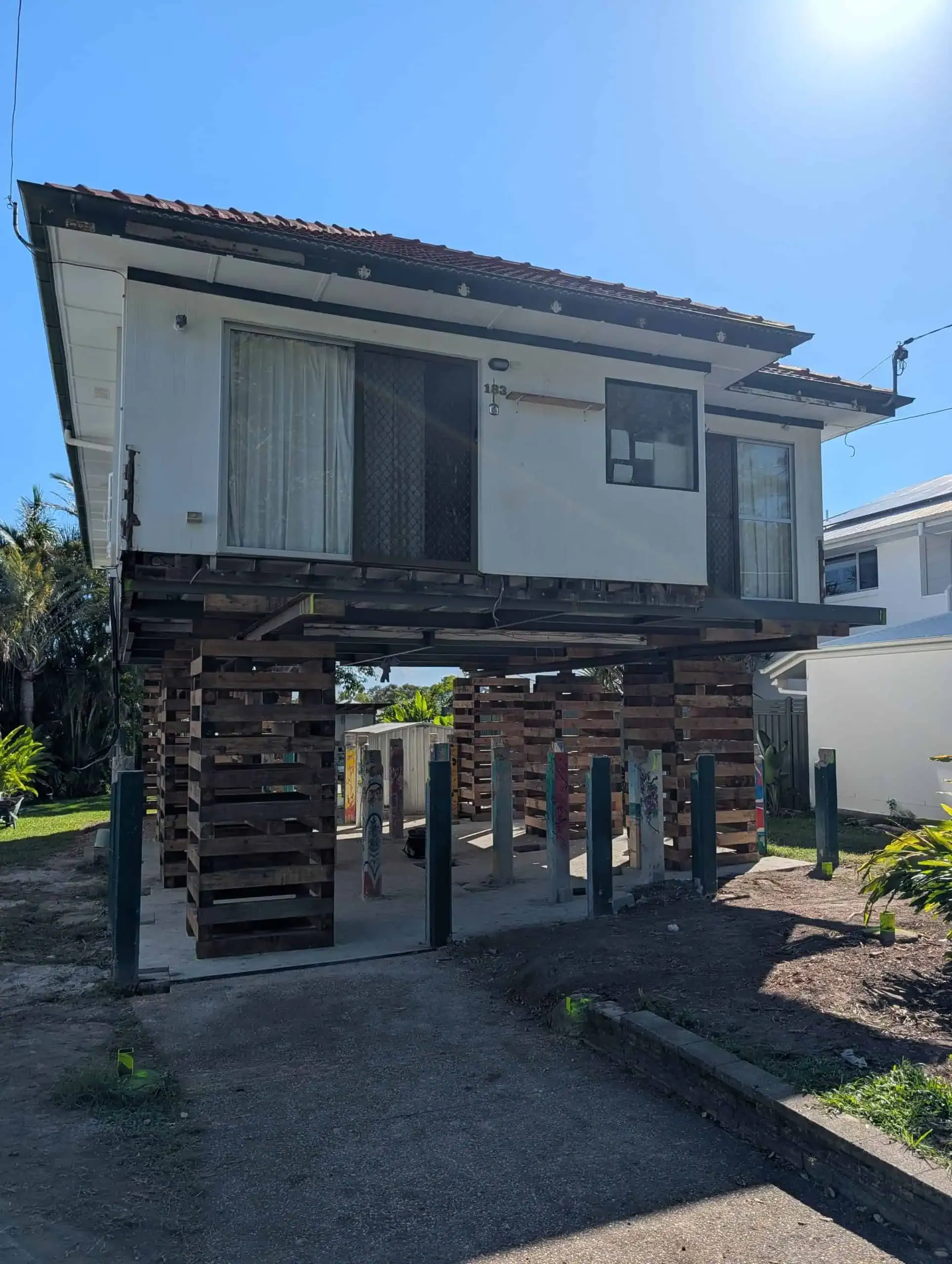 Stilted wooden house with a weathered exterior and stairs, raised above a gravel lot under bright blue sky