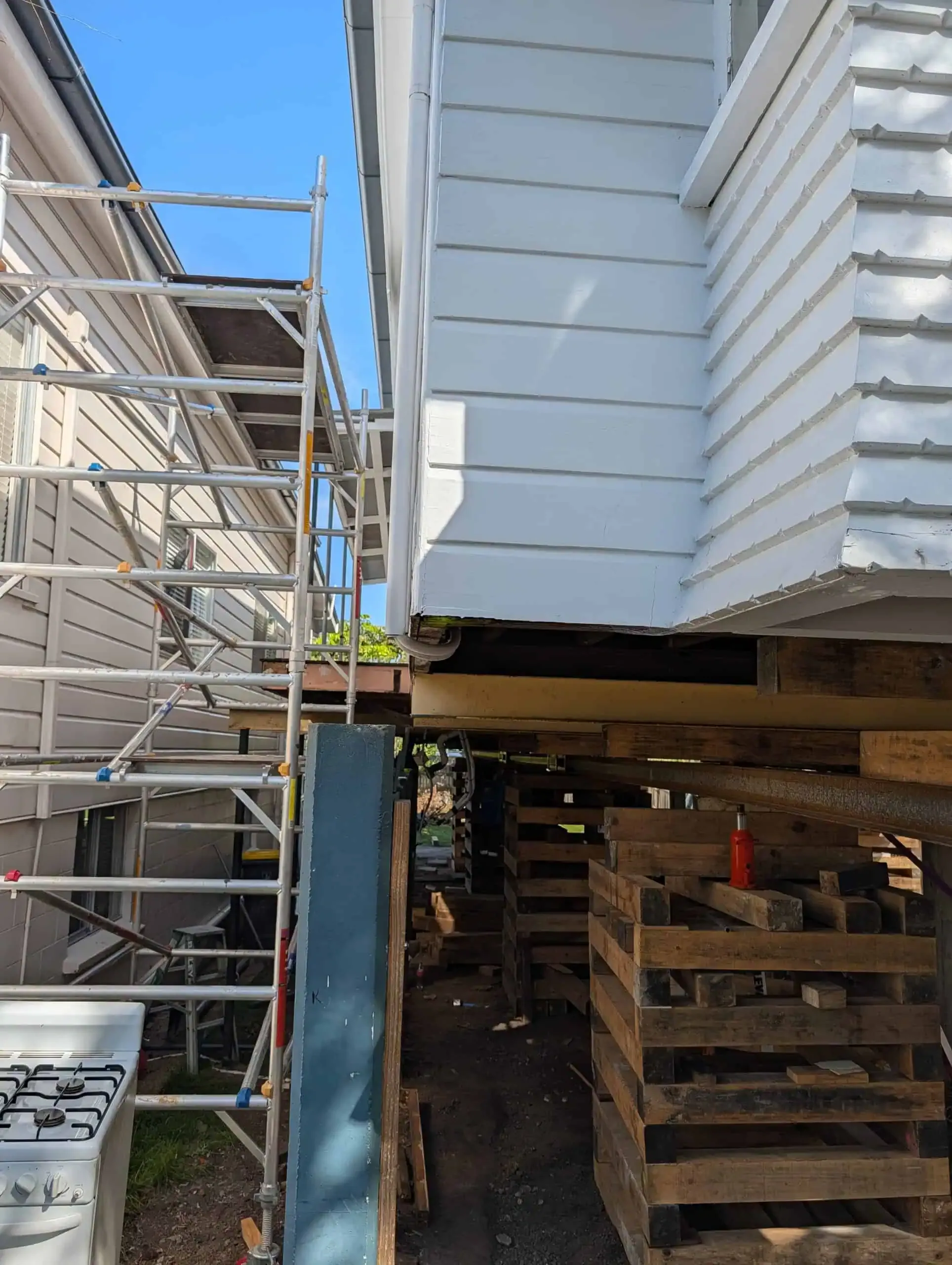 House raised on stilts beside scaffolding and a narrow gap, with stacked lumber underneath.