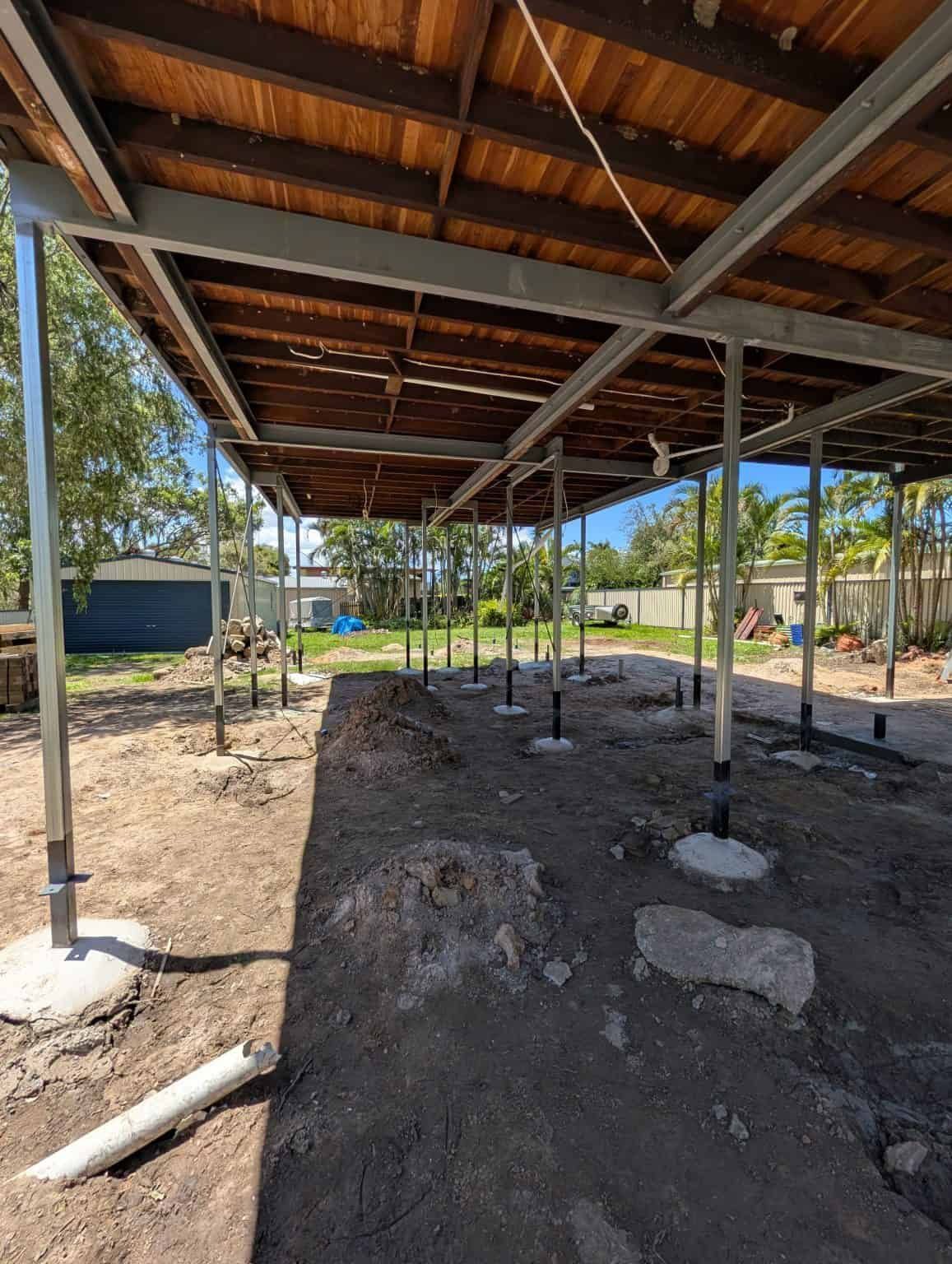 Partially built pavilion with roof beams and metal posts over a dirt floor outdoors