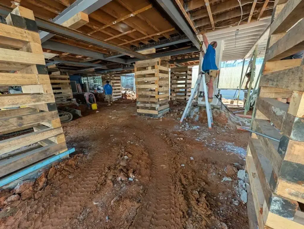 Construction site interior with stacked pallets, muddy floor, and a worker on a ladder under exposed beams