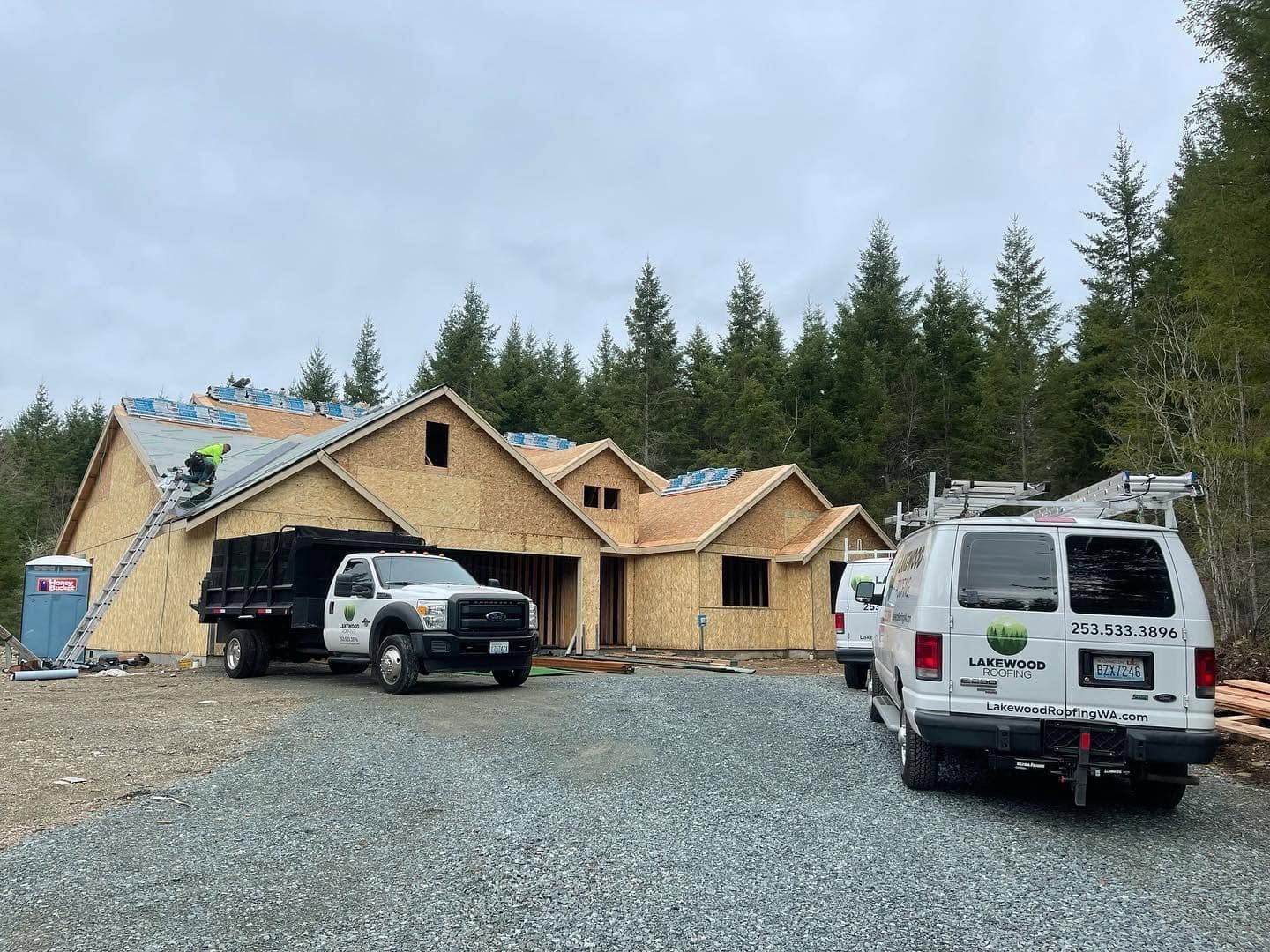 Partially built house with two trucks on a gravel driveway, surrounded by trees under a cloudy sky