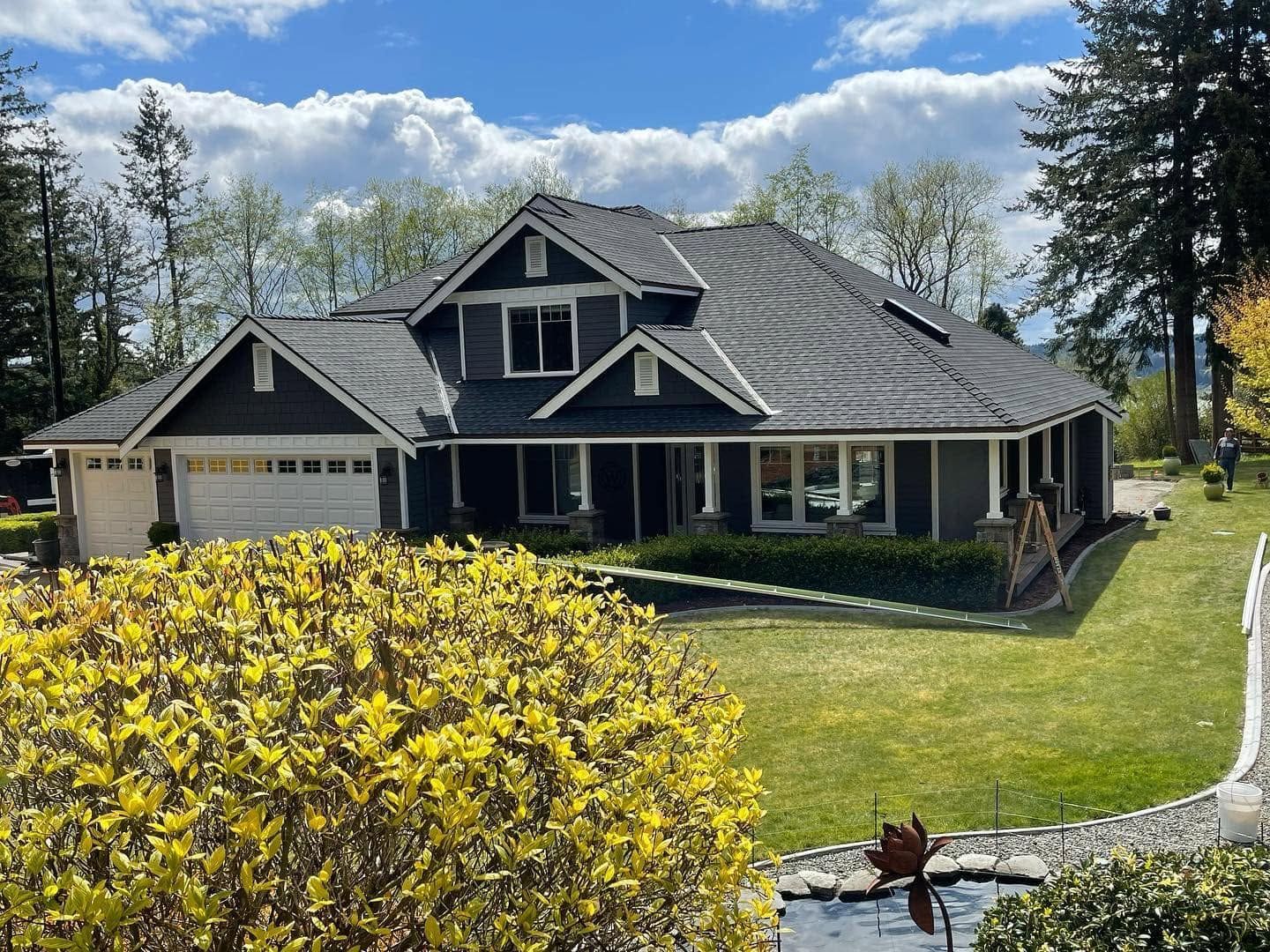 Suburban house with dark roof, white trim, and a yellow shrub in the foreground on a sunny day