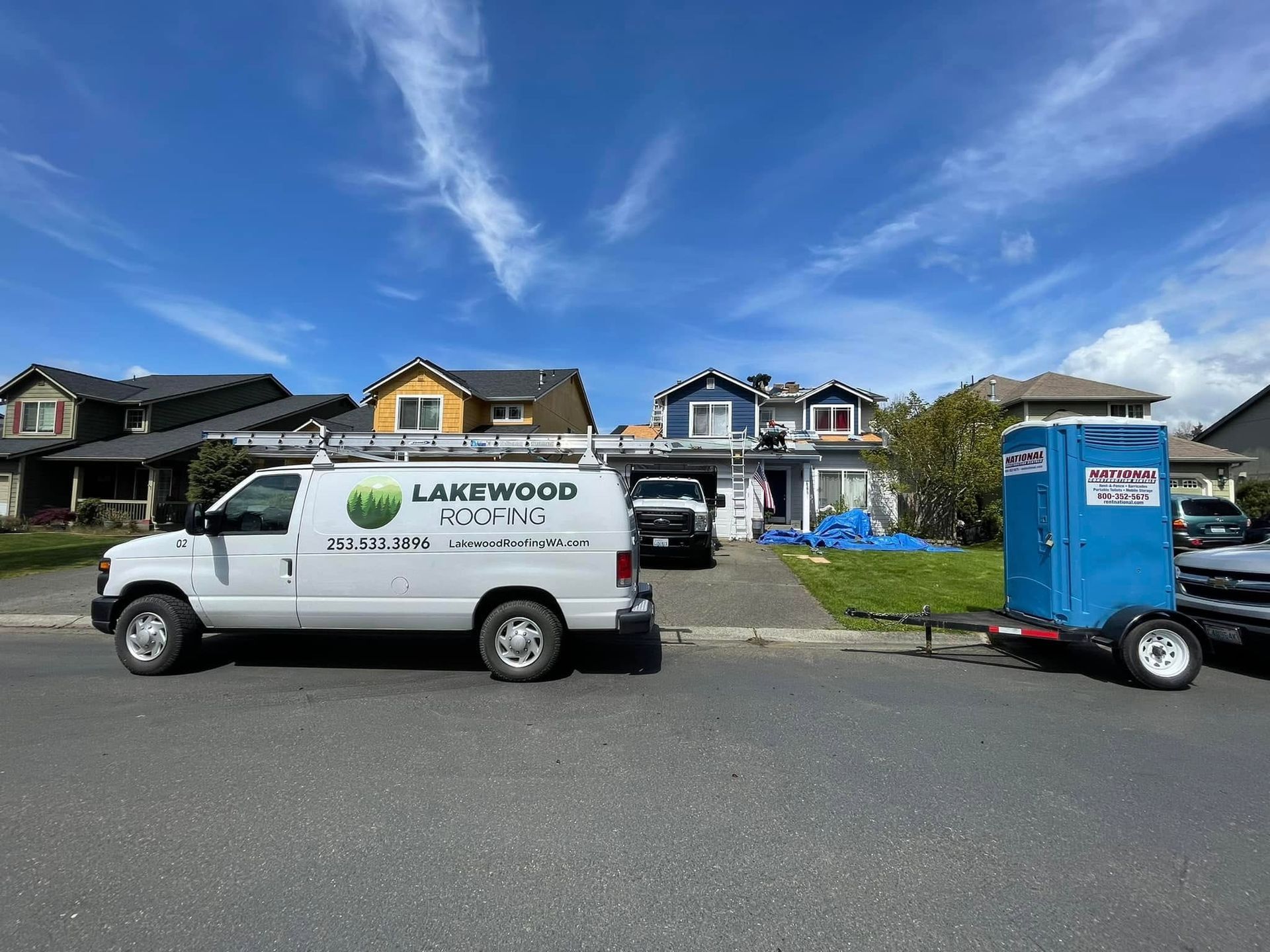 White Lakewood Roofing van and blue portable toilet trailer parked on a residential street under a blue sky