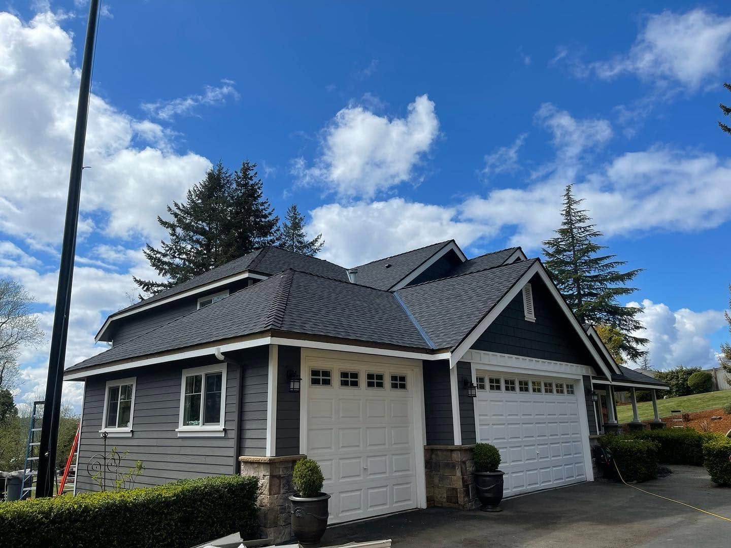 Gray two-car garage with dark roof under a blue sky with clouds and tall trees nearby