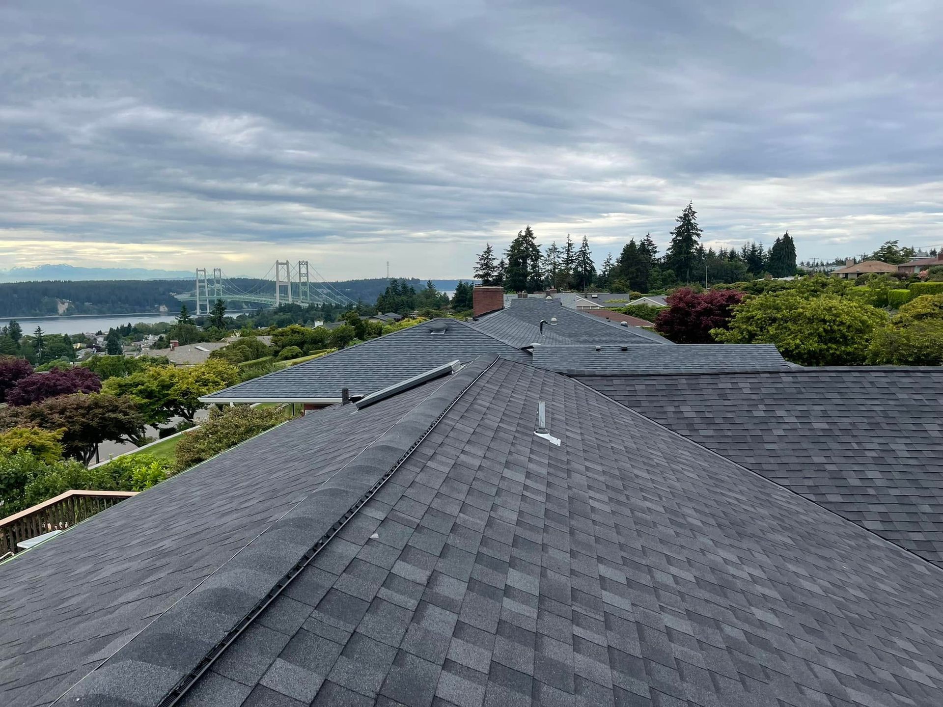 Rooftop view of gray shingle roofs overlooking trees, a cloudy sky, and distant city buildings