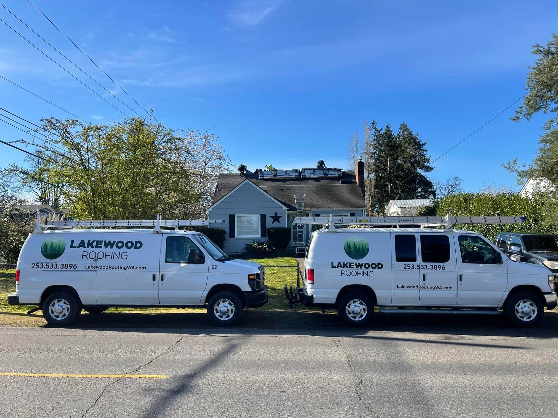 Two white Lakewood Roofing vans parked on a street in front of houses under a blue sky