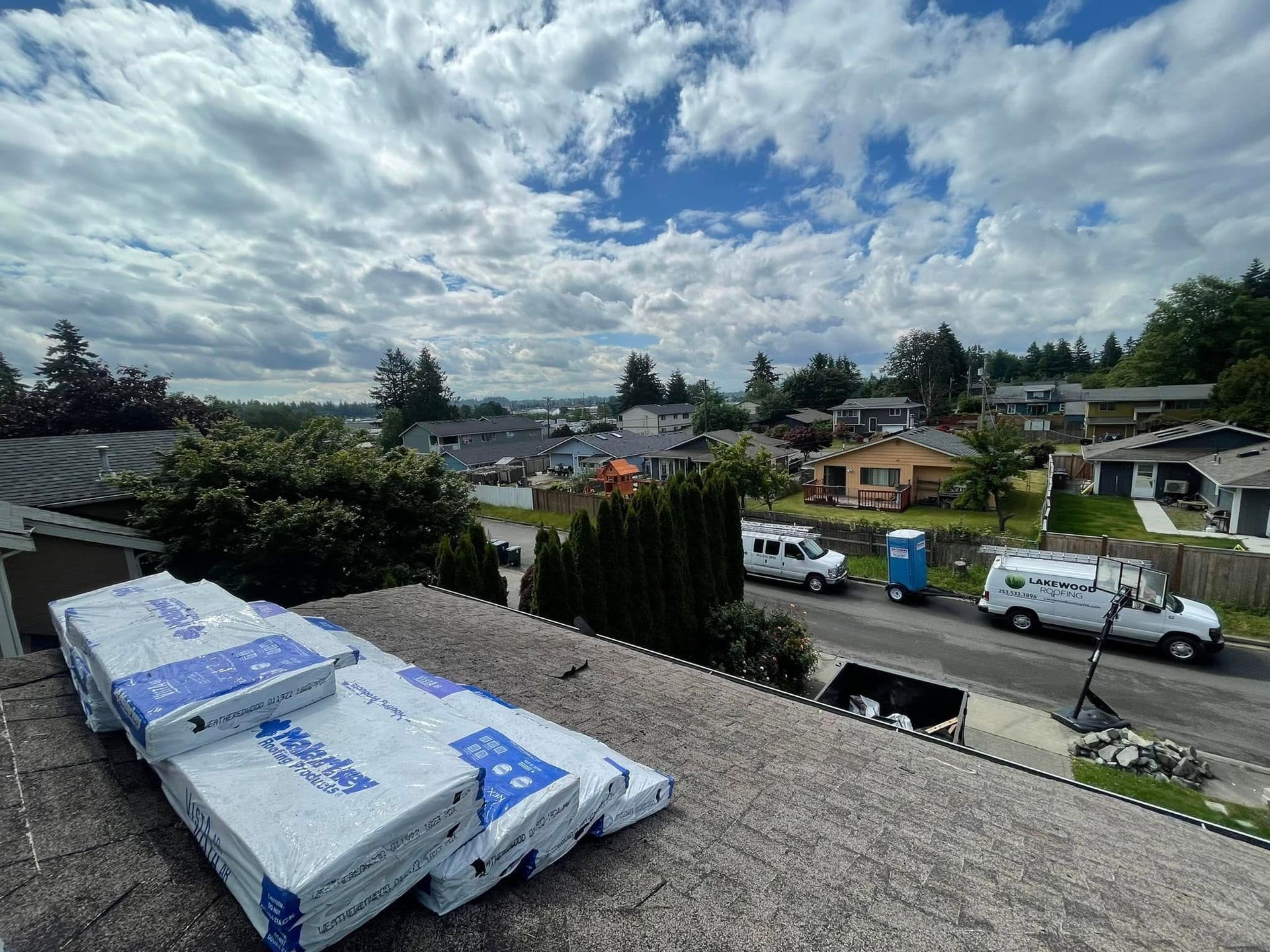 Rooftop view of a suburban neighborhood with stacked roofing materials, parked trucks, and a cloudy sky.