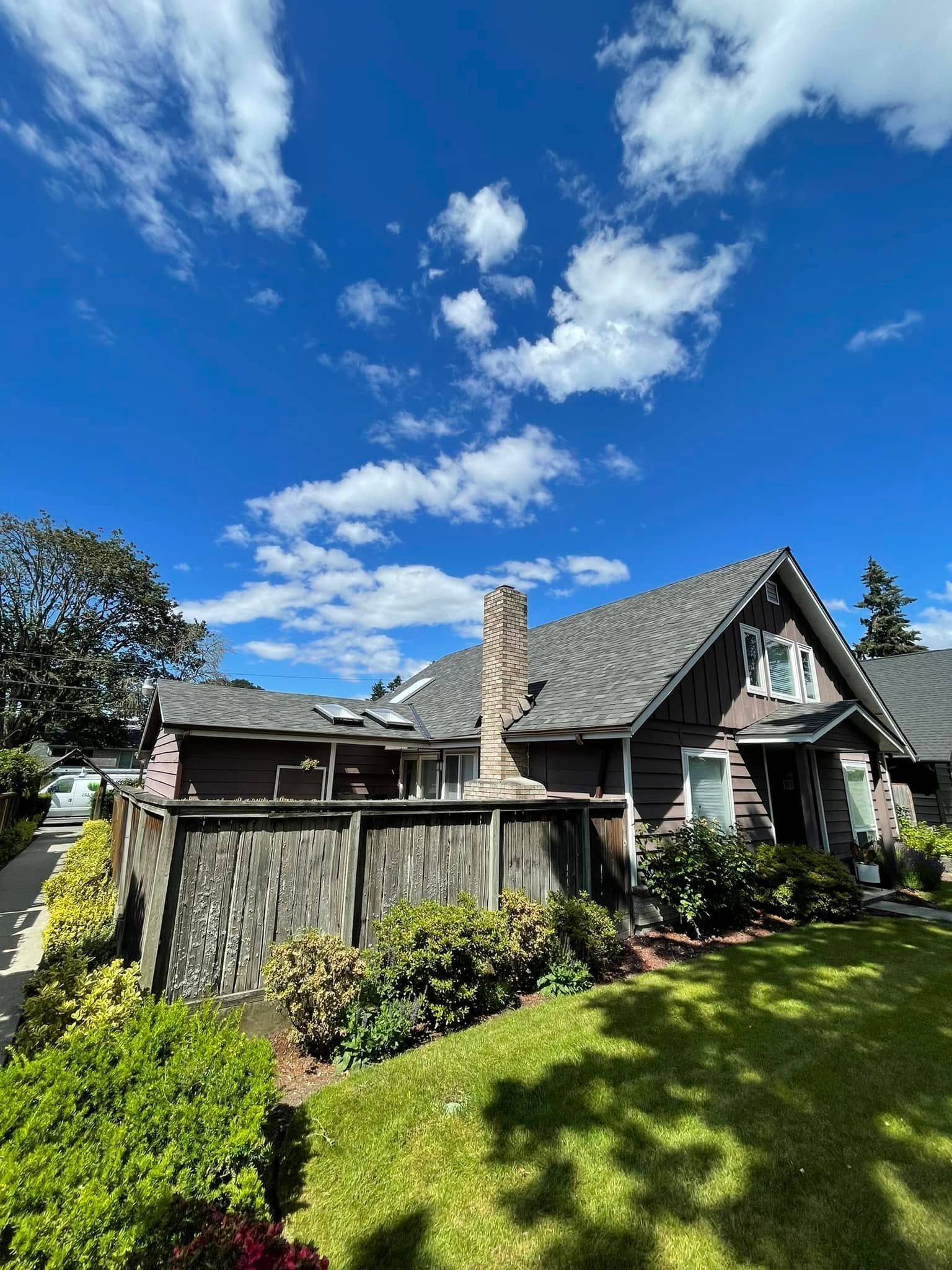 Suburban house with a stone chimney, wooden fence, green lawn, and blue sky with scattered clouds