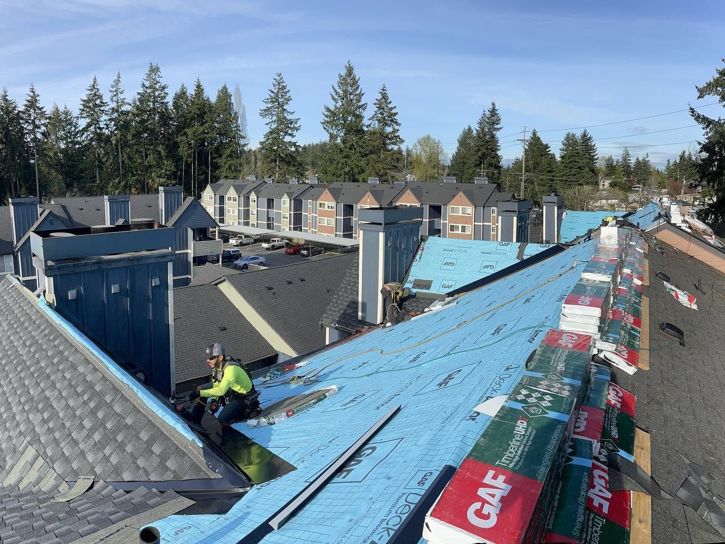 Workers on blue underlayment under repair with plywood and materials, viewed over neighboring houses and trees