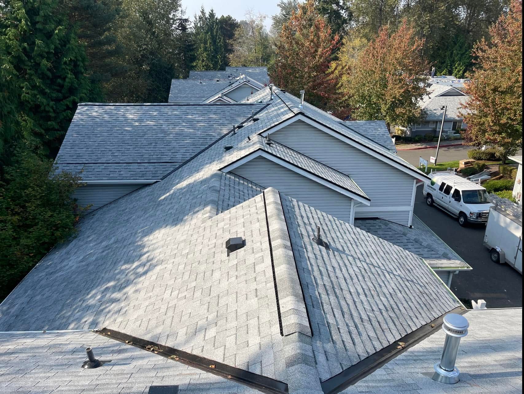Rooftops of a white house with gray shingles, surrounded by trees and parked vehicles.