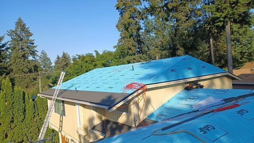 House roof under repair with bright blue tarps and red safety markers, surrounded by tall trees