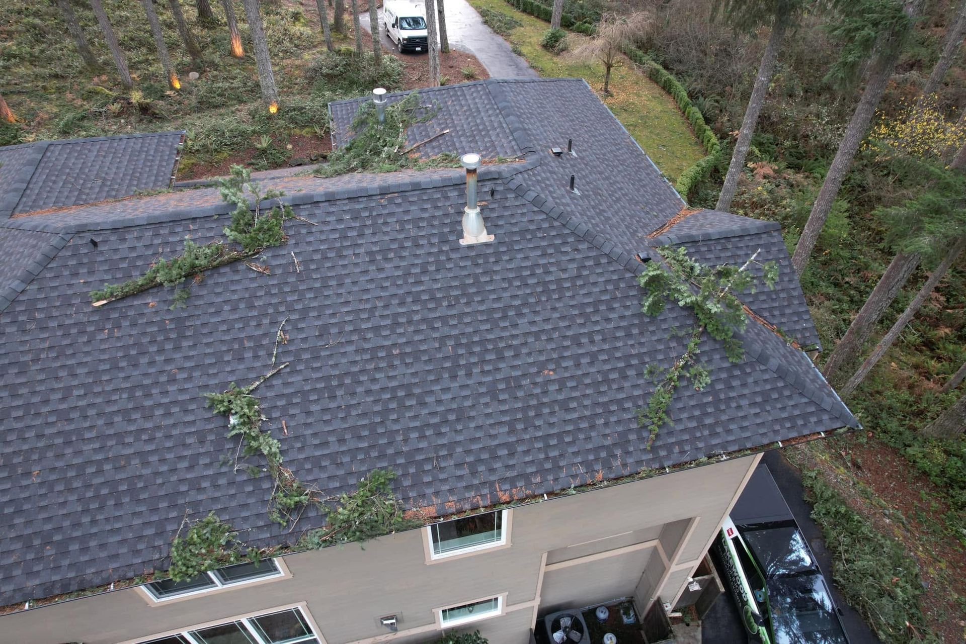Aerial view of a gray shingled roof with branches and debris, beside a wooded driveway and parked cars.