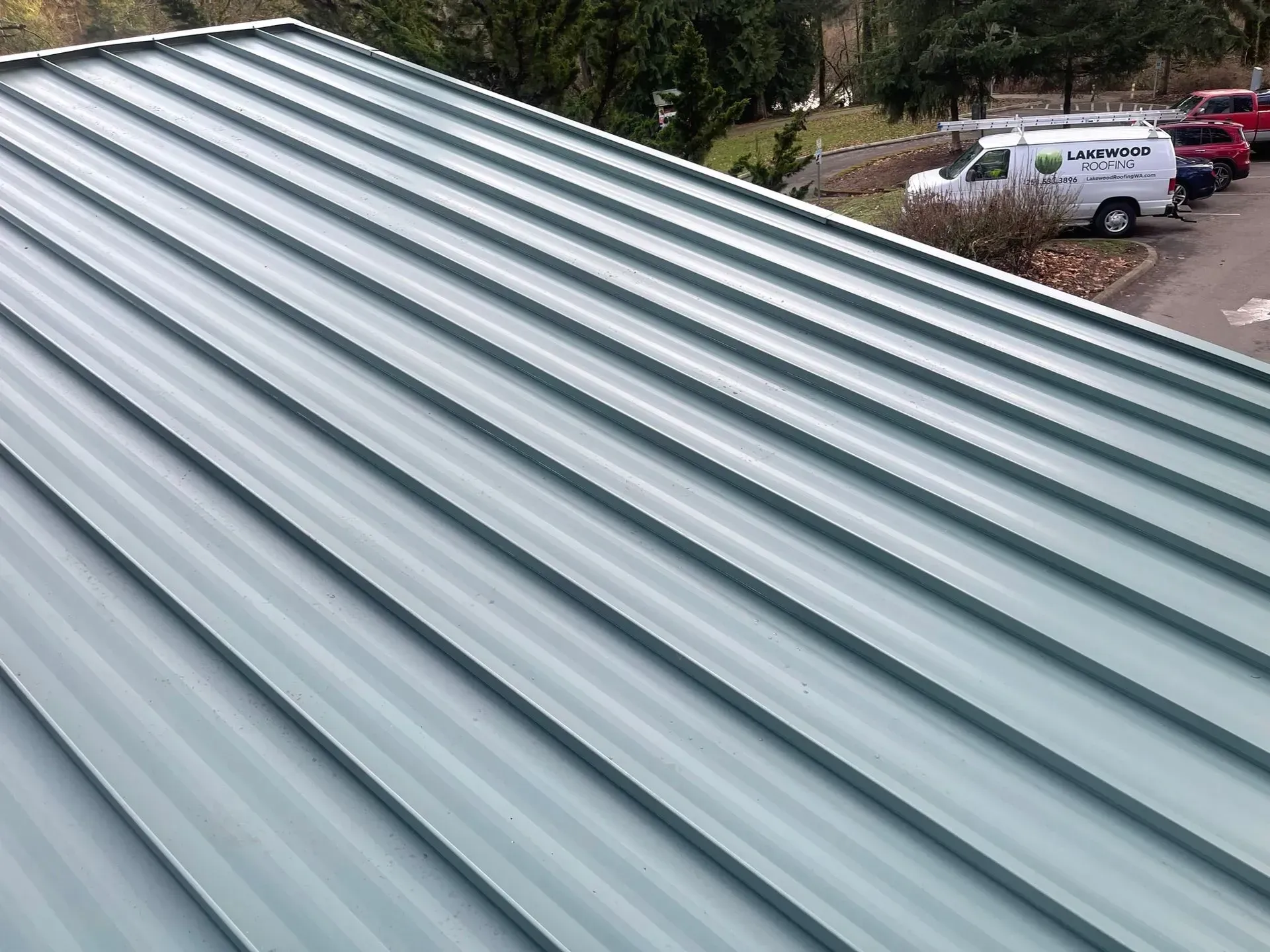 Close-up of a light gray corrugated metal roof with parked cars and trees in the background