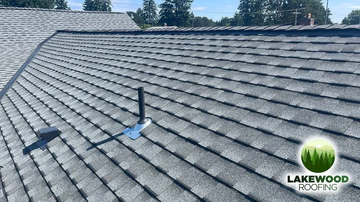 Gray shingle roof with a black vent pipe under a clear sky, Lakewood Roofing logo at bottom right