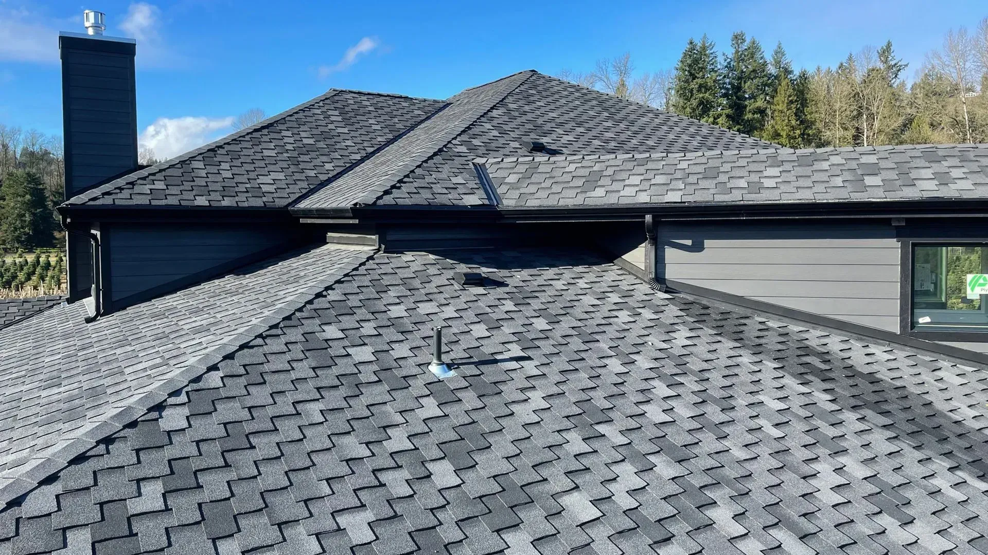 Dark gray shingled residential roof with a chimney and skylights under a clear blue sky