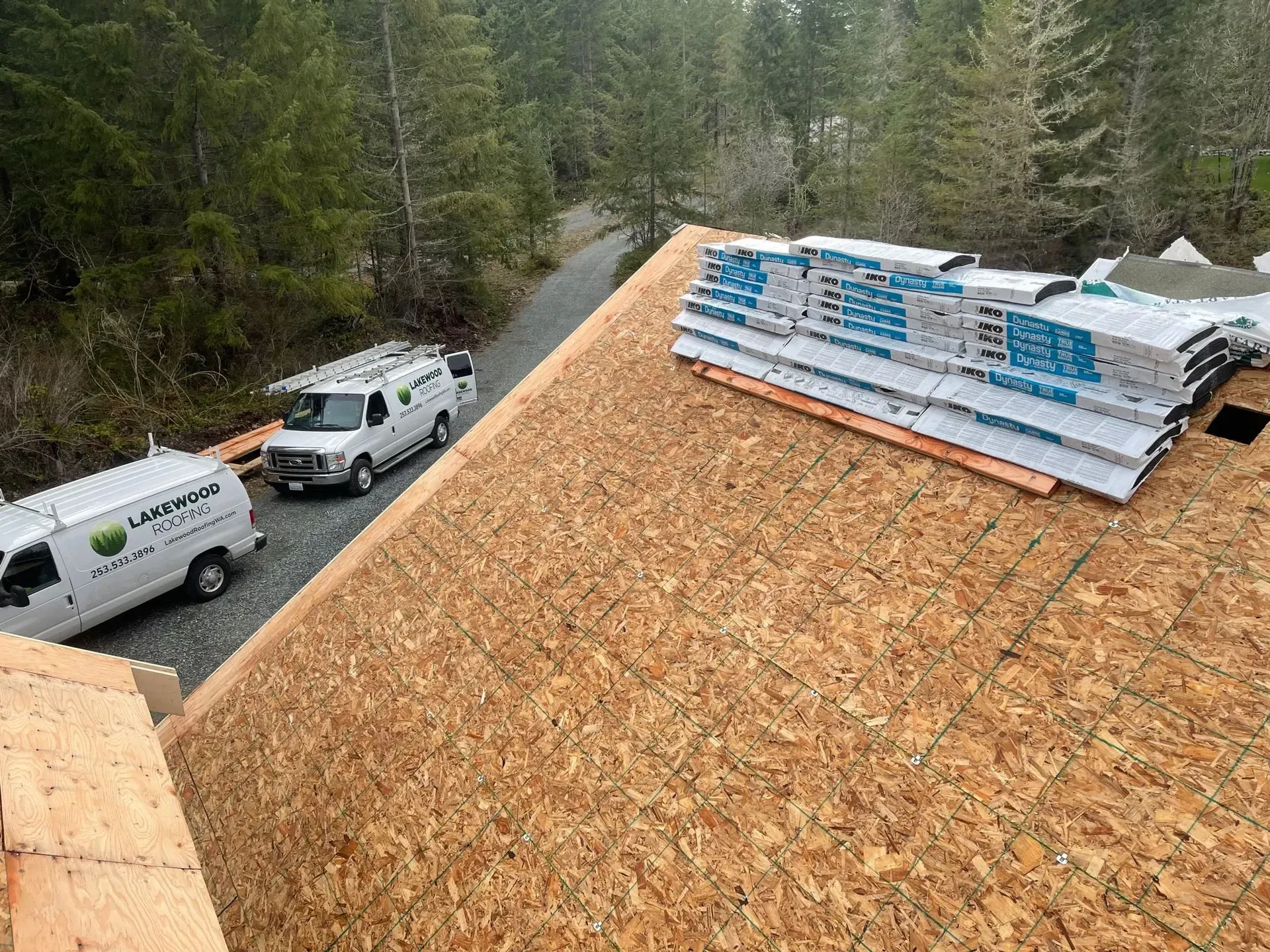 Roofers working on a house roof with lumber in a wooded area, with service vans parked below