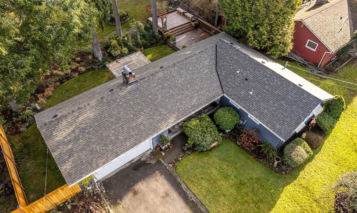 Aerial view of a single-story house with a gray roof, surrounded by trees and a green yard.