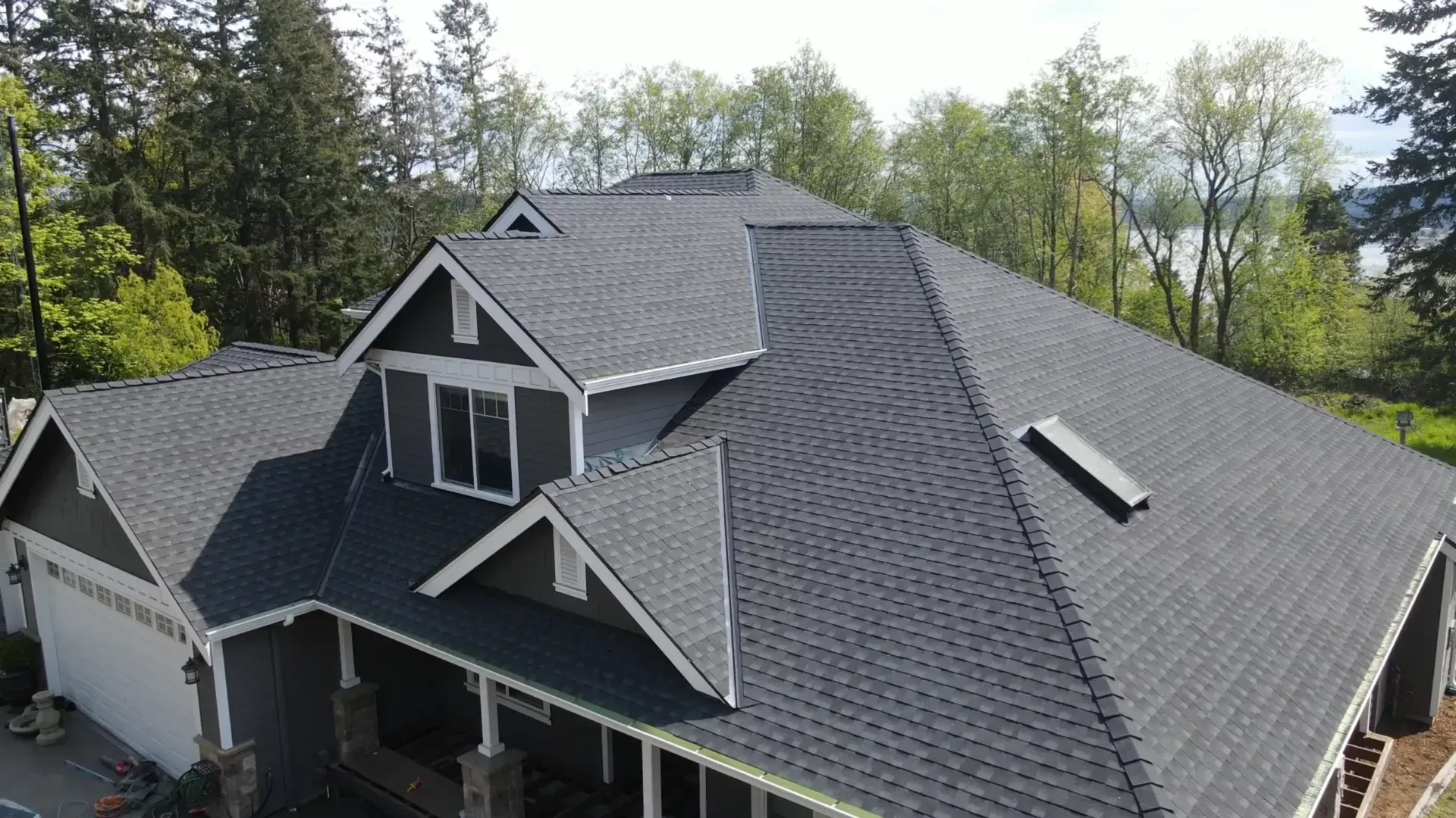 Aerial view of a house with dark gray shingle roof and dormers, surrounded by trees.