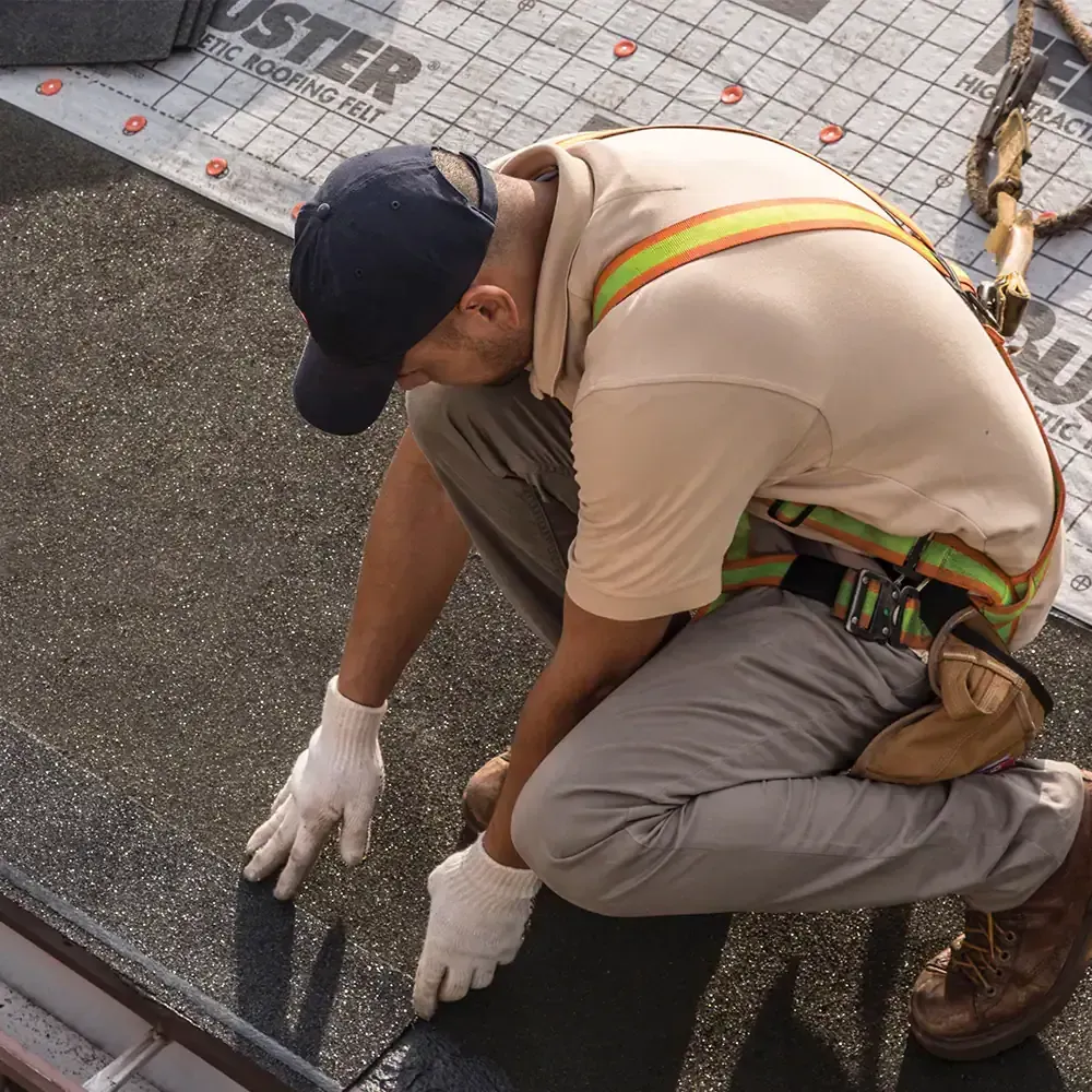 Worker in tan shirt and gloves kneeling on a rooftop, applying material along a roof edge.