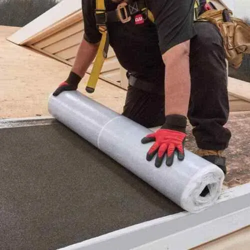 Worker rolling out gray roofing membrane on a flat roof, wearing gloves and harness.