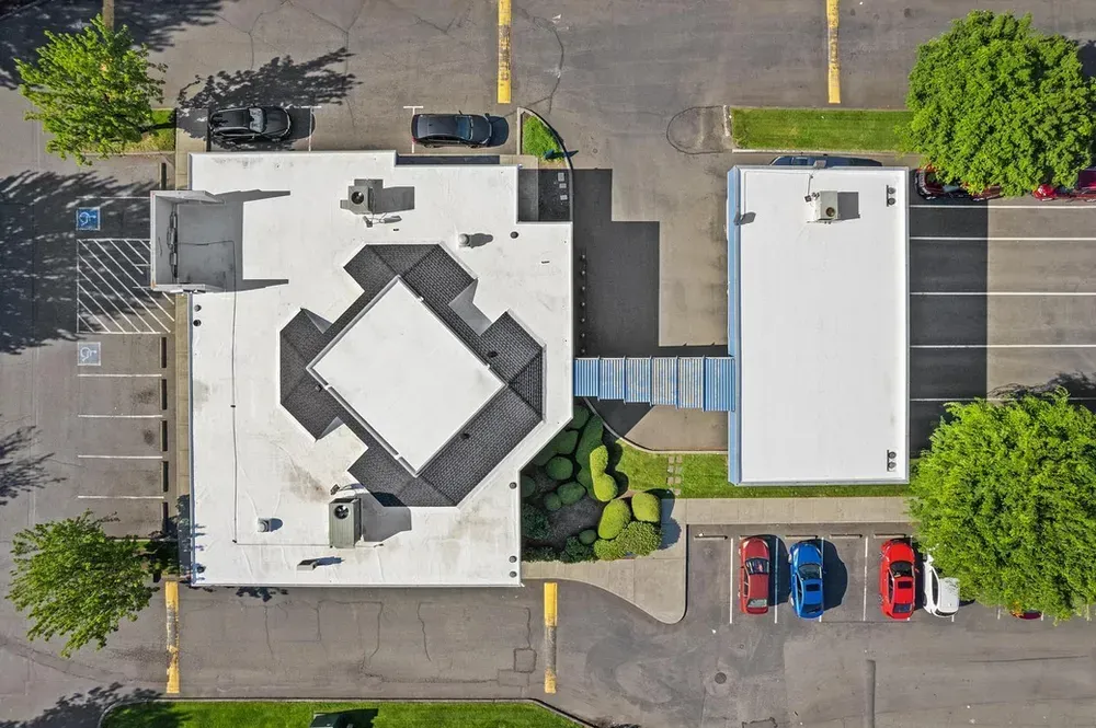 Aerial view of a building complex with white roofs, parking lots, trees, and three cars.