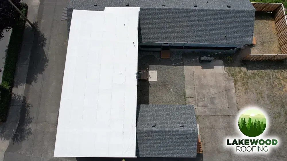 Aerial view of a roof with a white section and gray shingles, beside a yard and driveway.