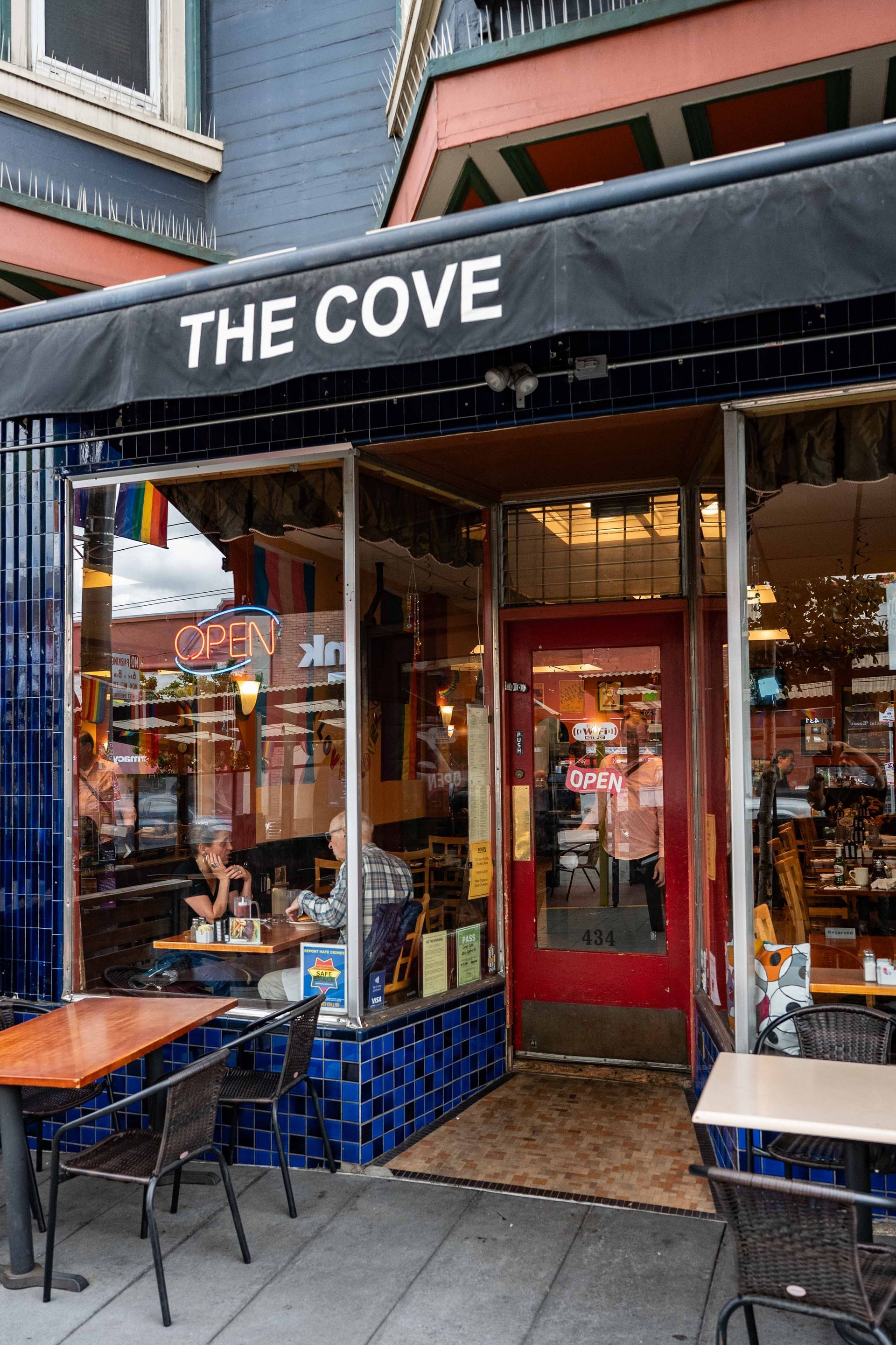 Storefront with black awning reading “THE COVE,” red door, and outdoor café seating