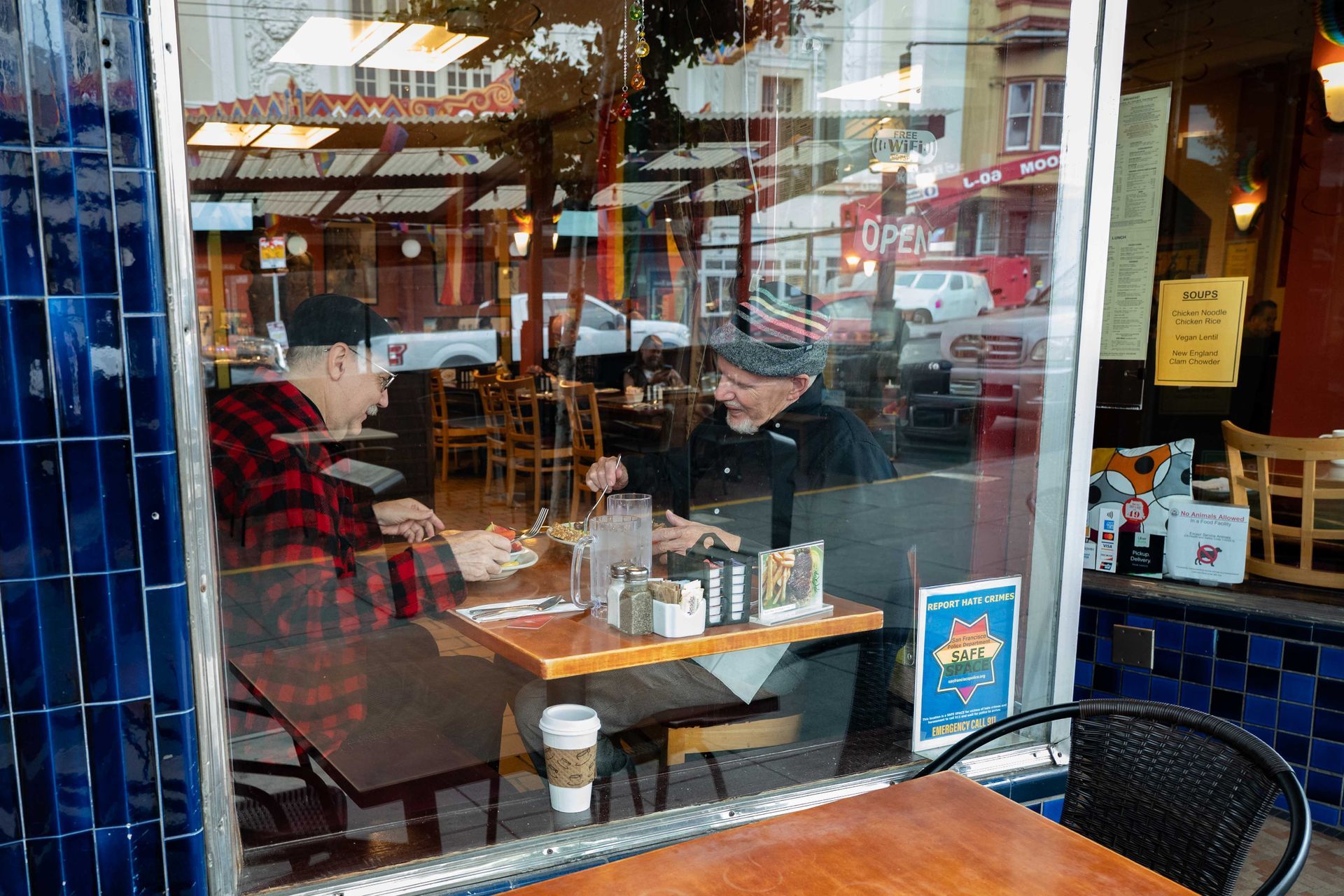 Two people sit at a café window table, with coffee cups and reflections of the street outside.