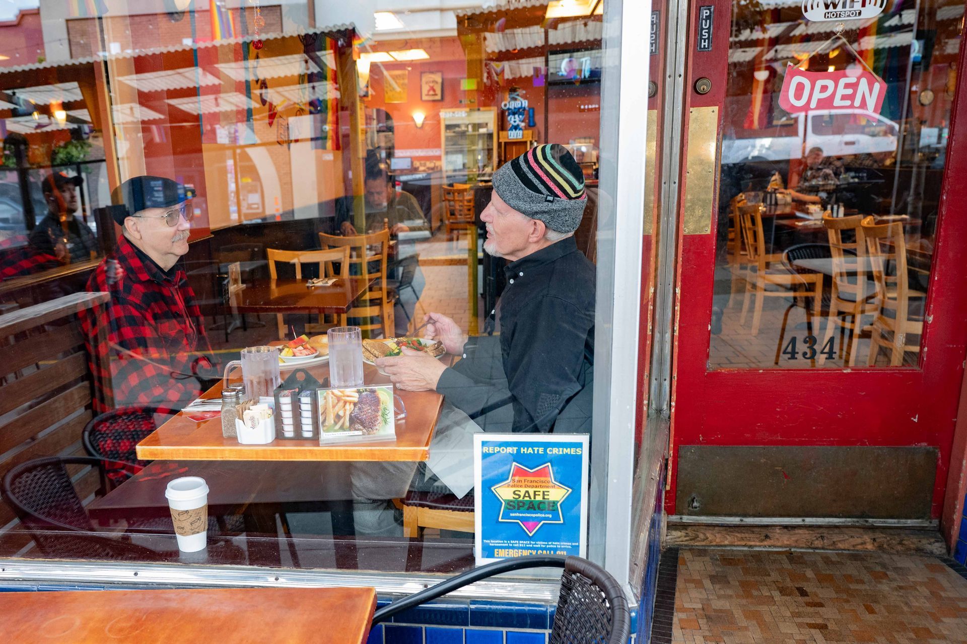 Two people talking at a café table by a red door with an “OPEN” sign