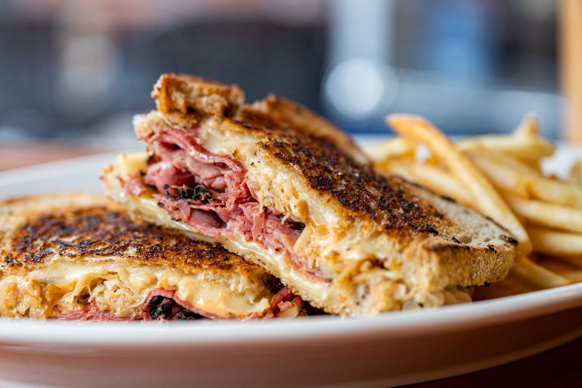 Grilled sandwich with melted cheese and sliced meat on a plate, with French fries in the background