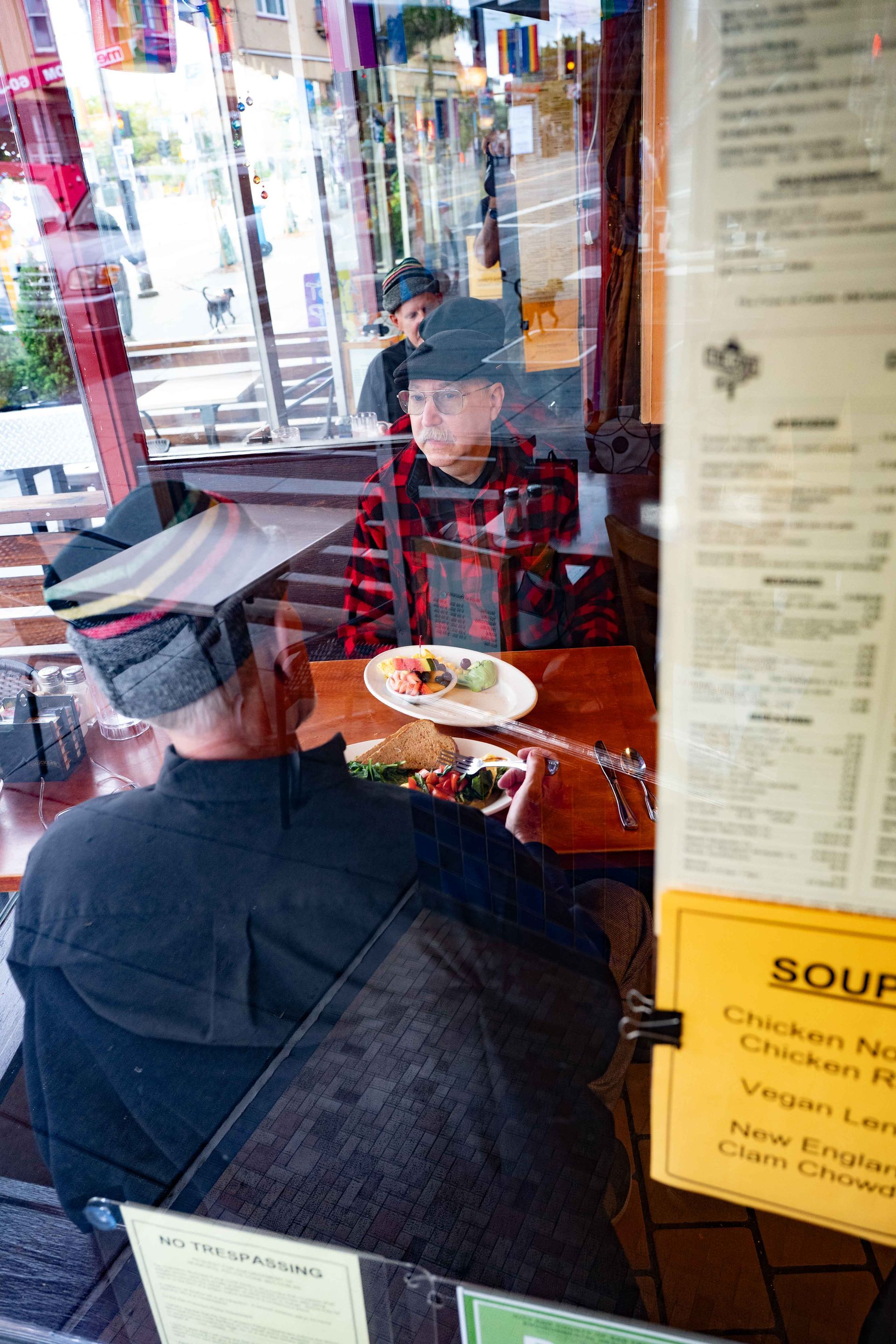 Two people seated at a café table, viewed through a window, with a menu posted on the right.