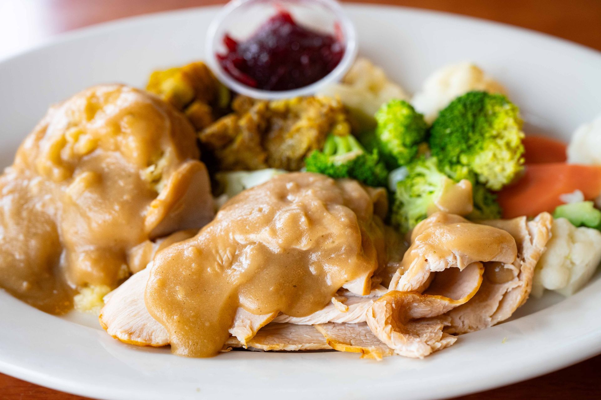 Plate of sliced turkey with gravy, mashed potatoes, broccoli, carrots, stuffing, and cranberry sauce