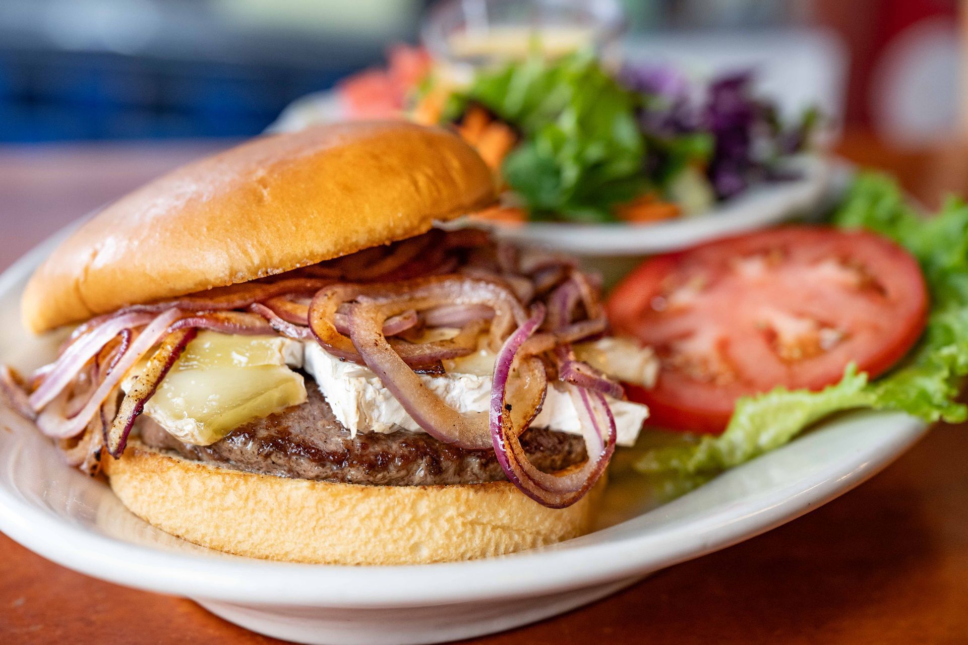 Cheeseburger with caramelized onions on a bun, served with tomato, lettuce, and salad on a white plate